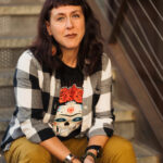 person with shoulder-length dark hair, a calavera t-shirt, black and white flannel, and green pants sitting on concrete steps near a metal railing. They are looking into the camera with a half smile.