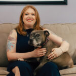 Tattooed middle-aged white woman with strawberry blonde hair and fair skin smiling while wearing a navy blue sleeveless dress, holding a tubby grey pitbull, and sitting on a couch.