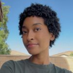 A close-up selfie of Trinity with short curly black hair, wearing a green shirt, smiling softly against a blue sky and natural background.