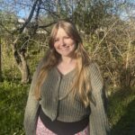 Smiling young woman with long blonde hair in a green sweater standing outdoors among trees.