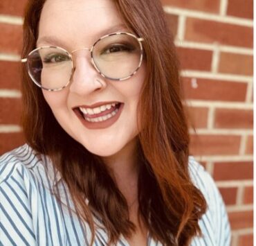 Image is a white woman with long red hair and glasses. She stands in front of a brick wall and wears a striped white and grey shirt. She's smiling.