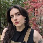 Jess has long brown hair and a collection of flower, bunny, and mushroom tattoos on their arm. They are wearing a pair of overalls and are standing behind a dark red Japanese maple tree.