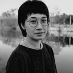 In a black and white image, a mixed race Korean person with short hair, round glasses, and a nose piercing is looking at the camera and smiling slightly. They are wearing a light t-shirt under a dark sweater and standing in front of a river, which is reflecting the trees.