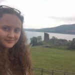 Image is of a white brunette woman with curly hair standing in front of a grassy landscape.