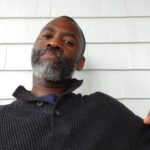 A Black man with a greying beard, in a grey sweater, sitting in front of a white wall, looking toward the viewer.
