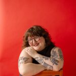 a feminine-presenting person sits, smiling, in a chair in front of a bright red backdrop. They have long, curly brown hair with bangs, clear and tortoise shell glasses, red lipstick, and a grey dress on.