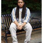 The photo is of a Black transmasculine man with long locs, a tan and blue striped shirt, tan pants, and red-brown loafers. He is sitting on a park bench with his hands clasped in front of him.