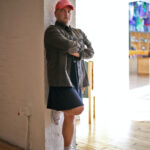 Nonbinary person with a hat, jacket, and skirt leaning agains a column.
