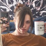 A mirror selfie of awhite woman with brown hair holding up a cat mug.