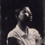 Black-and-white photo of a person, covered in heavy shadows, with light skin, a dark buzzcut, and facial piercings wearing a sleeveless white button-up, right cheek turned to the camera and looking up to the top right corner at an off-camera flash while reaching their right arm across their body and holding their left shoulder.