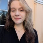 A woman with long, blonde and brown hair smiles into the camera. She is wearing a dark blue, button-up long sleeve and a gold necklace.