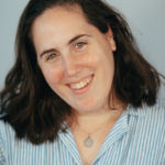 Livia, a white woman with dark brown hair and green eyes, smiles at the camera while wearing a blue and white striped shirt.