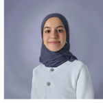 Headshot of brown woman of Middle Eastern/North African descent against a bluish-lavender background. She wears a greyish navy hijab (headscarf), silver earrings, and a white jacket with silver buttons.