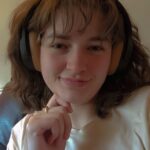 A white person with shoulder-length brown hair wearing black headphones and a white tee shirt smiles at the camera.