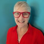 a smiling woman with short grey hair wearing red glasses and a red blouse and black pants against a teal wall
