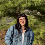 Headshot of Meghan Pinkston in front of a tree, smiling with hands in jacket pockets.