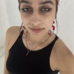Victoria is a woman with olive skin and dark brown hair that has been pulled back. She has black eyeliner on, is wearing long earrings that are clear and red, 3 chain necklaces, a small red horn charm, and is in a black sleeveless shirt. She is standing against a white background.