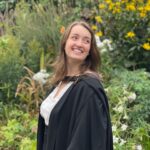 Headshot of Kaitlin Sandmann smiling wearing a black robe in front of flowers