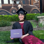 A woman with black hair sits outside in front of a green bush and sepia wall art. She is in her graduation cap and gown wearing a burgundy dress, holding an Emerson College diploma case.